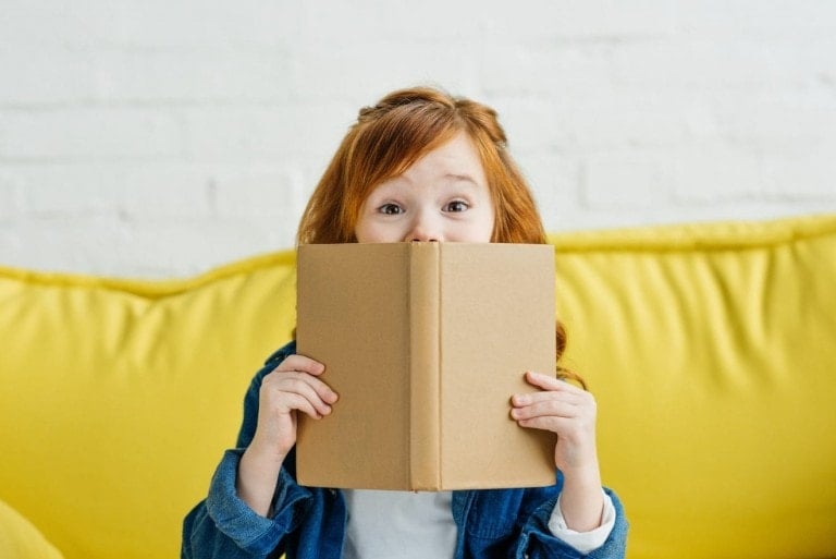 A young child with red hair is holding a brown book up to their face, with only their eyes visible over the top. They are sitting on a yellow couch with a white brick wall in the background. The child is wearing a blue jacket and a white shirt, deeply engrossed in one of the best children's books.
