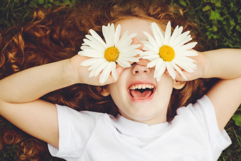 A child with red curly hair and a white shirt is lying on the grass, smiling widely. They are holding two large white daisies over their eyes, enjoying one of the fun spring activities for kids.