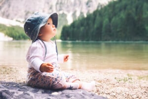 A baby sits on a blanket by the shore of a calm lake, wearing a blue sun hat, a light long-sleeved shirt, and floral pants. Against the backdrop of a forest and distant mountains under a clear sky, little River gazes up with a curious expression.