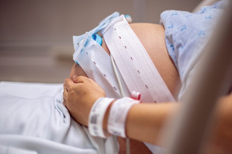 A close-up of a pregnant person's belly with medical monitoring belts attached, tracking early fetal heart rate decelerations. A hand with a hospital wristband is resting on the belly. The person is lying on a hospital bed, wearing a light blue hospital gown.