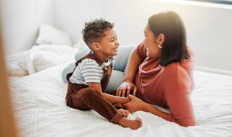 A woman and a young child are sitting on a bed, facing each other and smiling. The child is wearing brown overalls with a striped shirt, and the woman is wearing a pink top. This bright, well-lit room radiates warmth, fostering emotional development in children through shared moments of joy.