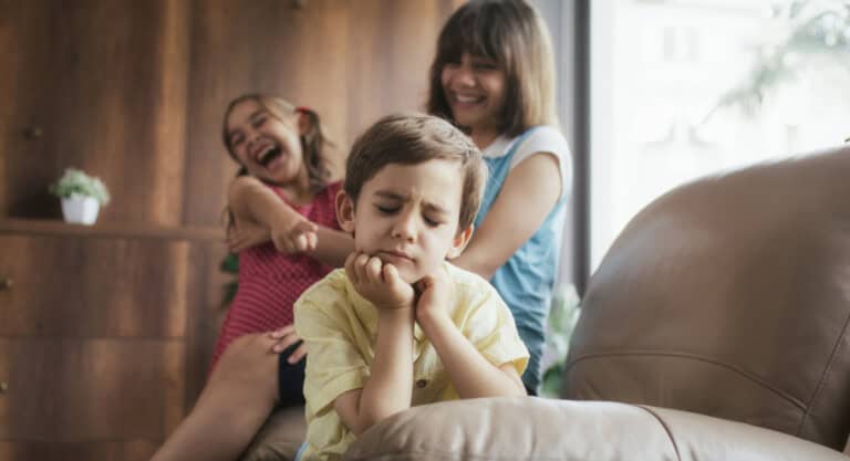 Three children are in a living room. In the foreground, a boy in a yellow shirt is sitting on a couch with a bored expression, resting his chin on his hands. In the background, two girls are laughing and one is pointing at the boy near a wooden cabinet—a reminder of things to never tease your child about.