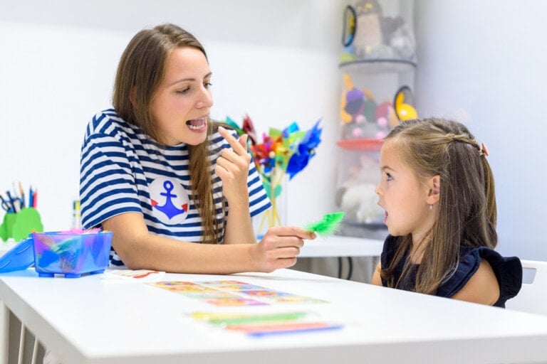 A woman in a striped shirt is holding a green feather and showing it to a young girl, engaging her interest. They are sitting at a white table with colorful items and drawings scattered on it in a bright room, where they might be discussing signs of speech and language delays.