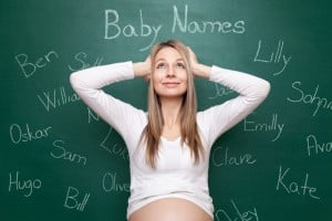 A pregnant woman in a white shirt holds her head with both hands and smiles while looking up. Behind her is a green chalkboard with various baby names written in different handwriting styles, including "Ben," "Lilly," "Sophia," "Kate," and "Hugo." She seems delighted as she searches for the perfect name.