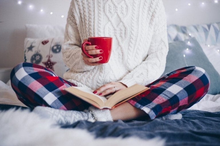 A person sits cross-legged on a bed, wearing a white knitted sweater and red and blue plaid pajama pants. They hold a red mug in one hand and have an open book on their lap, finding ways to practice mindfulness. The background includes pillows and string lights.