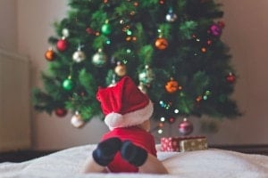 A baby celebrating their first Christmas, wearing a red Santa hat and outfit, lies on their stomach on a white blanket, mesmerized by a decorated Christmas tree adorned with colorful ornaments and lights. Two wrapped baby Christmas gifts sit nearby, completing the festive scene.