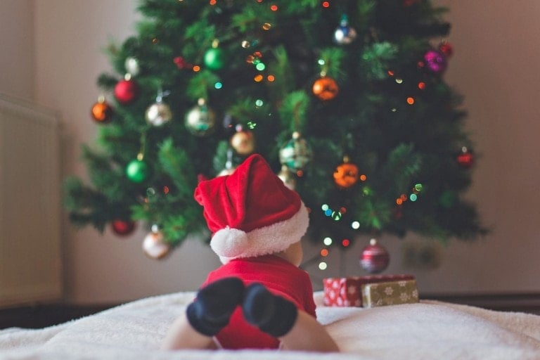 A baby celebrating their first Christmas, wearing a red Santa hat and outfit, lies on their stomach on a white blanket, mesmerized by a decorated Christmas tree adorned with colorful ornaments and lights. Two wrapped baby Christmas gifts sit nearby, completing the festive scene.