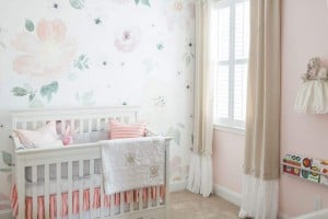 A beautifully organized baby nursery with light pink and white decor features a crib with a quilt and pillows, floral wall decals, a window with beige curtains, a small shelf with children's books, and a doll hanging on the wall. The room is carpeted and well lit by natural light.