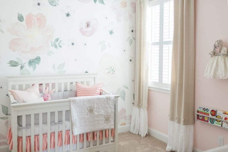 A beautifully organized baby nursery with light pink and white decor features a crib with a quilt and pillows, floral wall decals, a window with beige curtains, a small shelf with children's books, and a doll hanging on the wall. The room is carpeted and well lit by natural light.