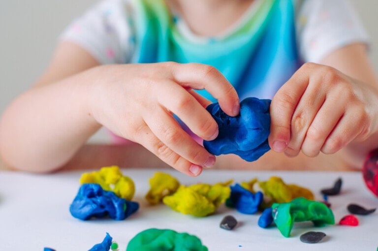 A child's hands are seen molding blue homemade playdough while various other colors of playdough are scattered on a white surface. The child is wearing a multicolored shirt.