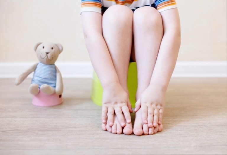 A child sitting on a green potty chair with their feet flat on the floor and hands touching their toes, demonstrating an ideal posture for boys' potty training. A stuffed teddy bear wearing a striped outfit is positioned on a pink stool nearby, set against a plain wall and wooden floor.