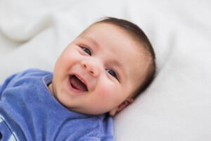A baby with dark hair lies on its back on a white blanket, wearing a blue shirt, and smiling with its mouth open and eyes wide. The background is out of focus, highlighting the little one's joyful expression as if enjoying the melody of beautiful Latin baby names being whispered.