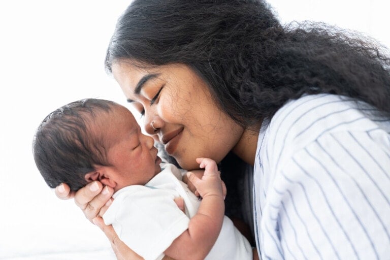 A woman with long, curly hair gently holds a newborn baby close to her face, sharing a tender moment with their eyes closed. The baby, wearing a white onesie, is surrounded by softly lit white tones. Amidst this peaceful scene, one might wonder: how much does it cost to have a baby?