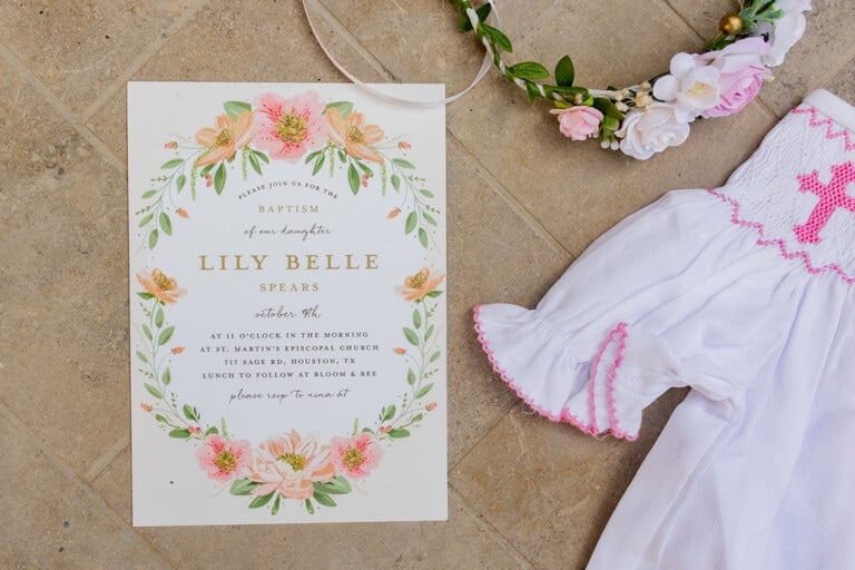 A baptism invitation for Lily Belle featuring floral decorations beside a floral headpiece and white dress with pink trim. The invitation provides details of the baptism ceremony at St. Martin's Episcopal Church in Houston, TX, with lunch to follow.