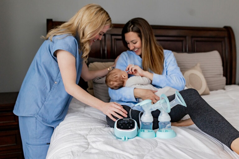 A woman in blue scrubs stands next to a smiling mother breastfeeding her baby on a bed. Beside them, on the white quilt with a brown headboard, is a Zomee Z2 double electric breast pump with bottles attached.