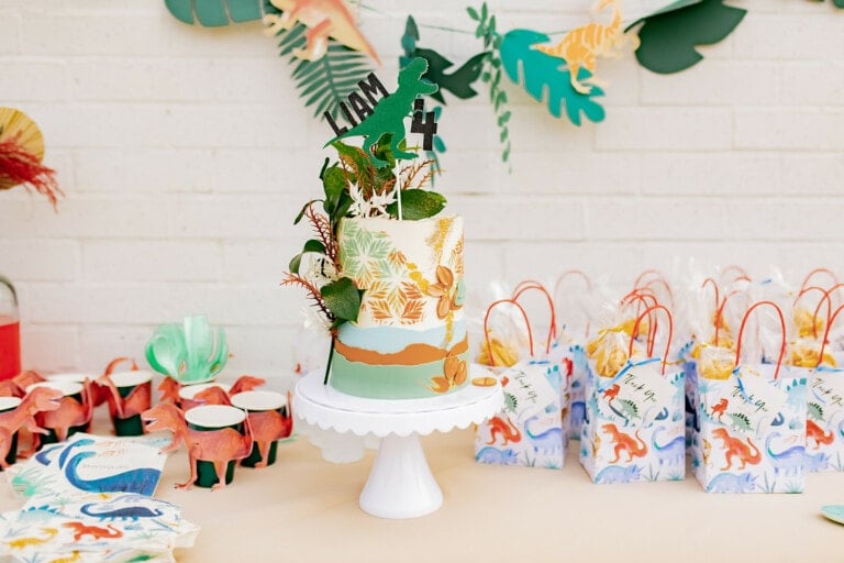 A white and green cake decorated with leaves and dinosaur figures sits on a white cake stand. Next to it are goodie bags with dinosaur designs and small red and green paper dinosaur figures. A Dinosaur Birthday Party banner hangs on the wall behind.