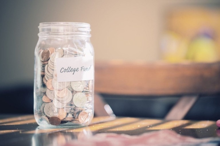A glass jar labeled "Child College Fund" is filled with coins and some dollar bills. The jar sits on a table with sunlight filtering in, casting shadows. Other blurred objects are visible in the background.