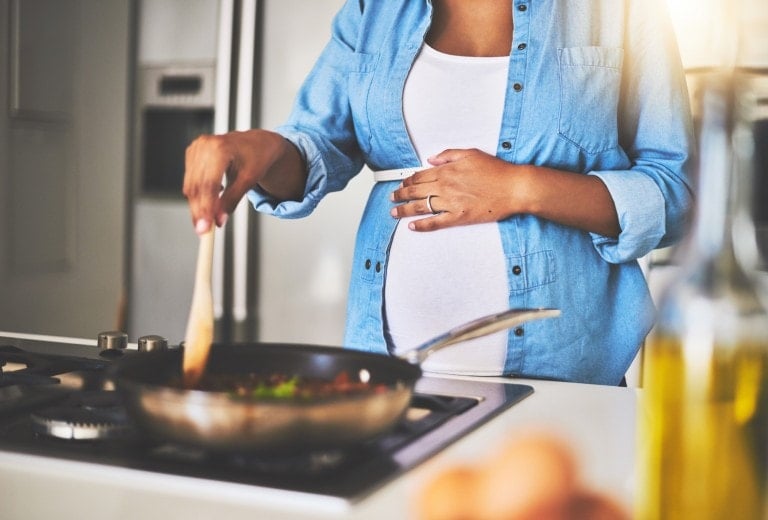 A pregnant woman stands in a kitchen, stirring food in a frying pan on a stovetop with a wooden spoon. She is wearing a white shirt and a blue denim jacket. Her other hand rests on her belly, perhaps contemplating third trimester recipes. Various kitchen items are visible in the foreground and background.