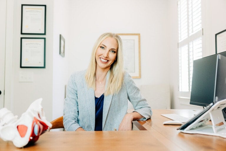 A smiling woman with long blonde hair is seated at a wooden desk in an office. She is wearing a gray blazer over a blue top. Behind her, certificates and framed documents are hung on the wall, showcasing her expertise in pelvic floor health. A computer and anatomical model are on the desk.