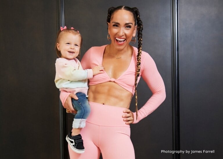 A person dressed in a pink long-sleeve crop top and leggings is holding a smiling toddler with a pink hair bow, embodying the spirit of "strong baby." The adult, reminiscent of Robin Arzón, smiles confidently and displays impressive abdominal muscles. The backdrop is black with "Photography by James Farrell" in the bottom right corner.
