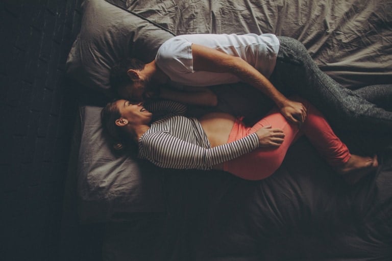 A couple lies on a bed, facing each other. The man is wearing a white t-shirt and gray pants, while the woman in a striped long-sleeve shirt and pink pants rests her hand on her pregnant belly. Both smile warmly, wrapped in love languages of pregnancy under a cozy gray blanket.