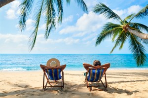 Two individuals are reclining on striped beach chairs under palm trees, facing a calm blue ocean with a clear sky. The person on the left is wearing a wide-brimmed hat, and both appear to be enjoying a kid-free vacation on the sandy beach, taking in the serene surroundings.