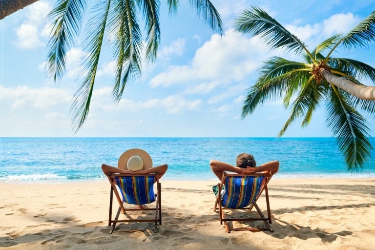 Two individuals are reclining on striped beach chairs under palm trees, facing a calm blue ocean with a clear sky. The person on the left is wearing a wide-brimmed hat, and both appear to be enjoying a kid-free vacation on the sandy beach, taking in the serene surroundings.