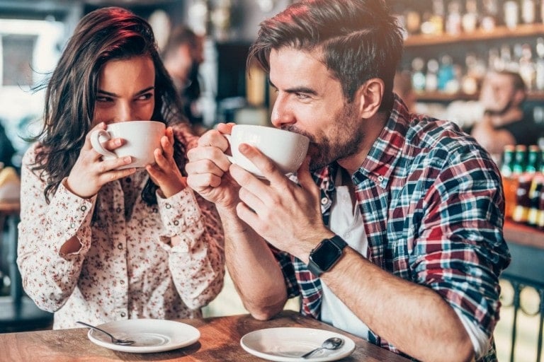 A woman and a man are seated at a cafe, sipping from large white mugs and smiling. The woman is wearing a floral blouse, and the man is wearing a plaid shirt and a smartwatch. There are empty saucers with spoons in front of them, illustrating one of the many low cost date night ideas. The background is blurred.