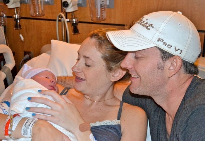 A woman in a hospital bed holds a newborn baby wrapped in a blanket, her face glowing as she shares this intimate birth story. A man wearing a white cap sits beside her, his arm around her shoulders as they both gaze at the baby and smile warmly. Medical equipment hums softly in the background.