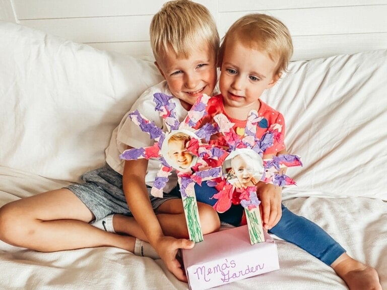 A young boy and girl sit on a white couch, each holding a colorful flower craft with photo centers, labeled "Nema's Garden". The boy is wearing a white shirt and gray shorts, and the girl is in a pink top and blue leggings. They both are smiling, showcasing their grandparent's day craft.