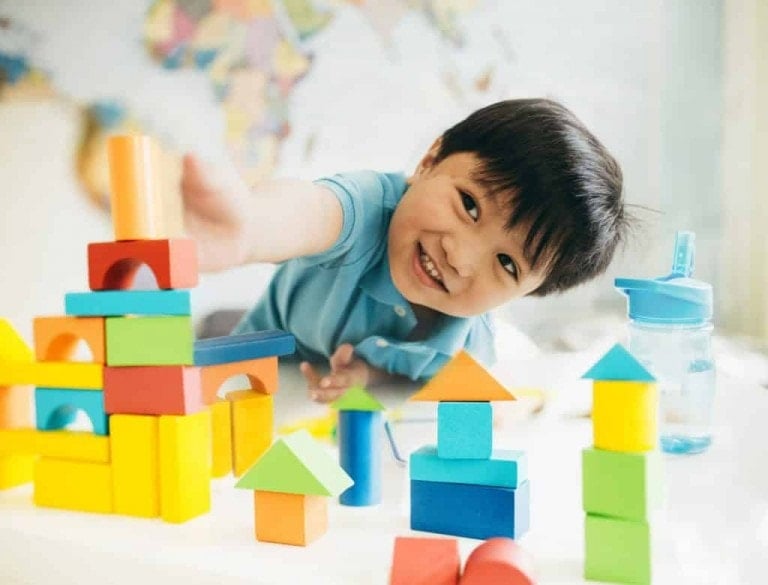 A young child wearing a blue shirt is smiling while engaging in purposeful play with colorful wooden blocks on a table. There is a blue water bottle next to the child. A world map is partially visible in the background.