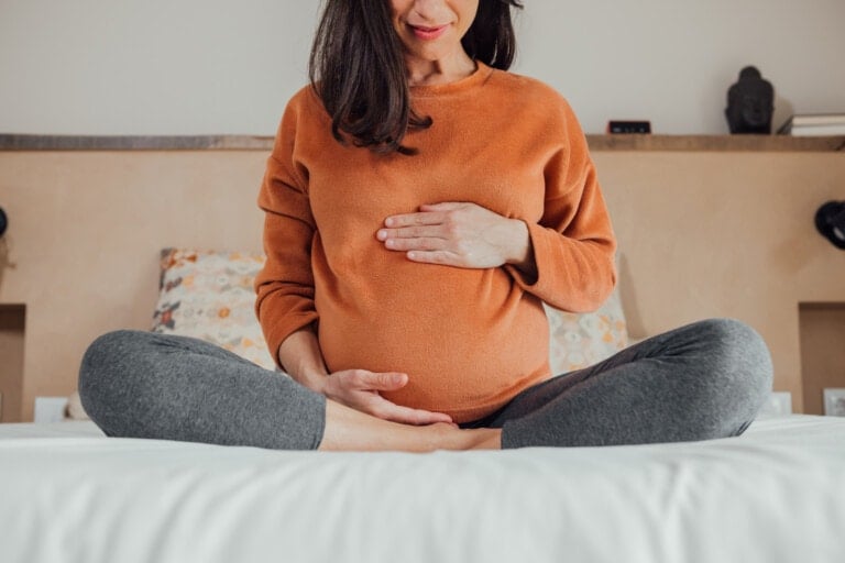 A pregnant woman sits cross-legged on a bed, wearing a rust-colored sweater and gray leggings. She gently holds her baby bump with both hands, embodying the spirit of mindful motherhood. The room has simple decor with patterned pillows and a shelf in the background.