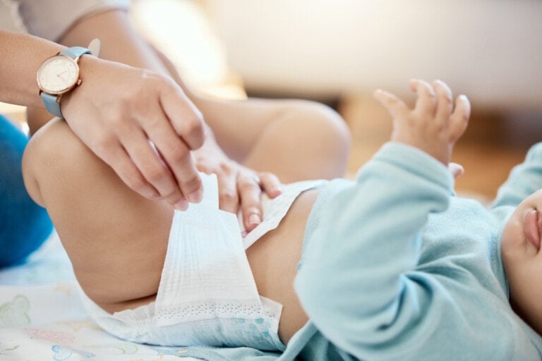 An adult with a wristwatch is changing a baby's diaper. The baby, dressed in a blue top, lies on its back with its legs slightly raised. The adult's hands are seen adjusting the waistband of the best diapers available. The scene is indoors in a well-lit room.