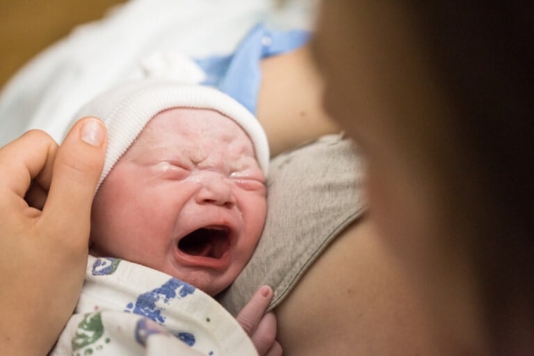 A newborn baby, with closed eyes and an open mouth, is crying while being held close to a person's chest. The baby, possibly having just received erythromycin eye ointment, is wearing a white cap and partially wrapped in a patterned blanket. A hand gently supports the baby's head.