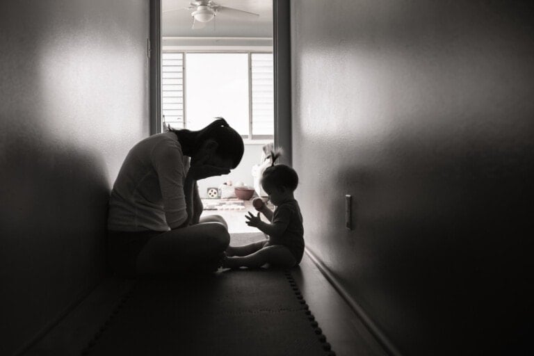 A black and white photo shows a woman and a baby sitting on the floor in a narrow hallway. The woman, displaying signs of mommy burnout, appears to be looking down while the baby holds up their hands. Light filters in from a window at the end of the hallway, illuminating the scene.