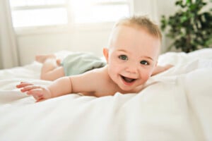 A baby with short light hair is lying on a bed on its stomach, looking at the camera with a wide smile. Soft, natural light from a window behind the baby bathes him in a gentle glow, while a green plant is partially visible in the background—a perfect moment for those considering boy names that start with F.