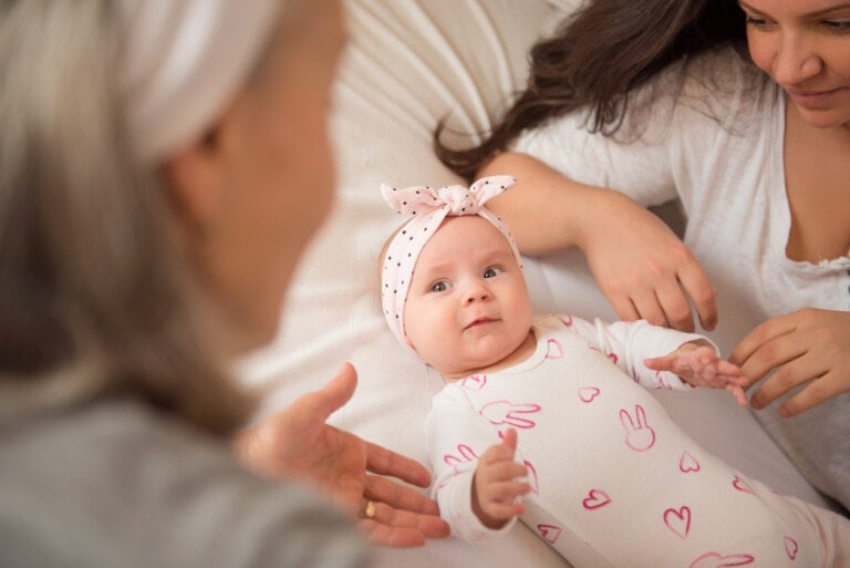 A baby dressed in a white onesie with pink bunny prints and wearing a pink headband lies on a bed. Two women, one with gray hair and another with dark hair, are looking at the baby and sharing bedtime routine tips while interacting with her.