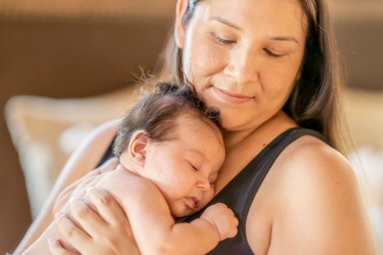 A woman with long hair is holding a sleeping baby close to her chest. The woman, rediscovering you after baby, is wearing a sleeveless top and looking down at the baby with a gentle expression. The background is blurred, focusing attention on the woman and the baby.