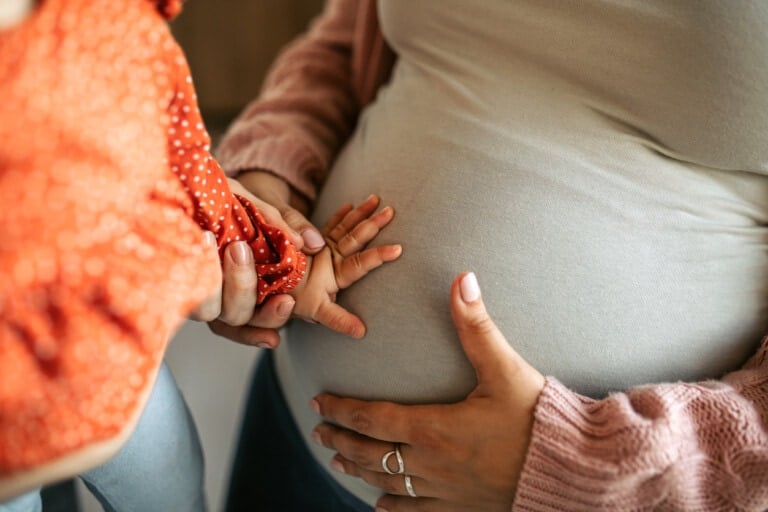 A close-up image of a pregnant person wearing a beige top and a pink cardigan, cradling their belly. A child in an orange and white polka-dot outfit is touching the pregnant belly with both hands, capturing the precious moment of going from 1-2 children.