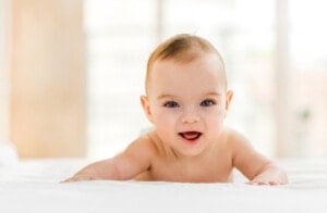 A baby with light skin and a small amount of hair lies on their stomach on a white surface, looking forward and smiling. The background is softly blurred, suggesting an indoor setting with natural light coming through windows. This serene scene captures the pure joy often associated with Ukrainian boy names like Andriy or Maksym.