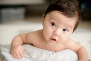 A baby with dark hair is lying on a light-colored pillow. The baby, perhaps destined for one of those charming Portuguese names, has large, expressive eyes and is looking slightly to the side with a curious expression. The background is blurred and neutral in color.