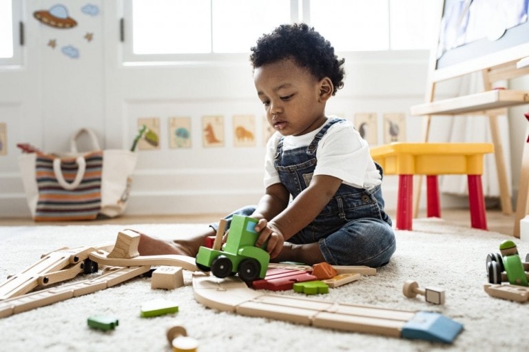 A toddler wearing a white shirt and denim overalls sits on a carpeted floor, engaging in unstructured play with wooden train tracks and a green toy train. The room is bright with natural light from the windows and has various toys and drawings in the background.