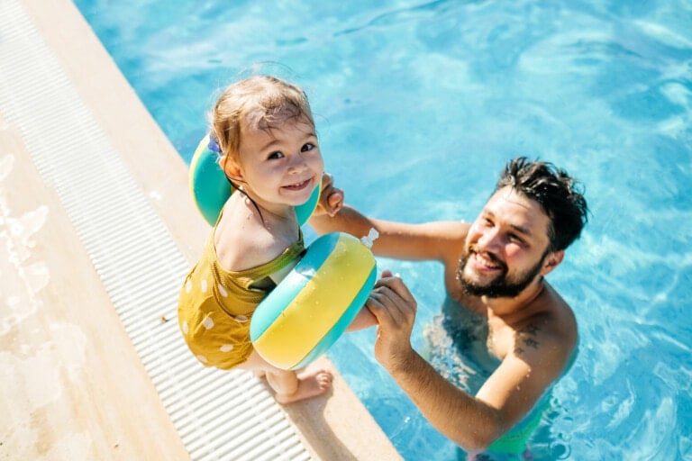 A man in a swimming pool holds up a smiling child who has inflatable arm floaties on, demonstrating excellent water safety. The child stands at the pool's edge, wearing a yellow swimsuit, while the man supports them from the water. The pool's clear blue water is visible in the background.
