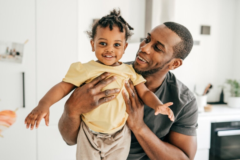 A man in a dark shirt is smiling and holding a baby dressed in a yellow shirt and beige pants. They are standing in a bright room with a white background. The baby is looking at the camera while the man looks lovingly at the baby, showcasing how dads are more involved today.