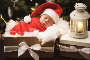A baby wrapped in a red blanket and wearing a Santa hat sleeps inside a gift box adorned with a white ribbon. The scene, perfect for holiday inspired baby names, includes soft white padding, a large nearby candle lantern, and blurred Christmas lights and decorations in the background.