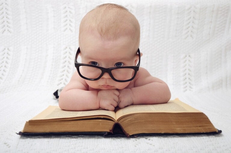 A baby with glasses, reminiscent of old-fashioned names like Henry or Edith, lies on its stomach with its elbows resting on an open book. The baby has a serious expression and is in front of a white, textured background.