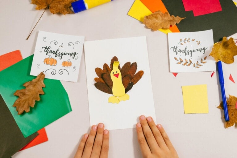 A person is creating a Thanksgiving-themed craft project. The project includes a yellow paper turkey with brown leaves as feathers glued to a white sheet of paper. Two Thanksgiving signs with decorative fonts and illustrations are positioned nearby, making this one of the most delightful Thanksgiving crafts for kids.