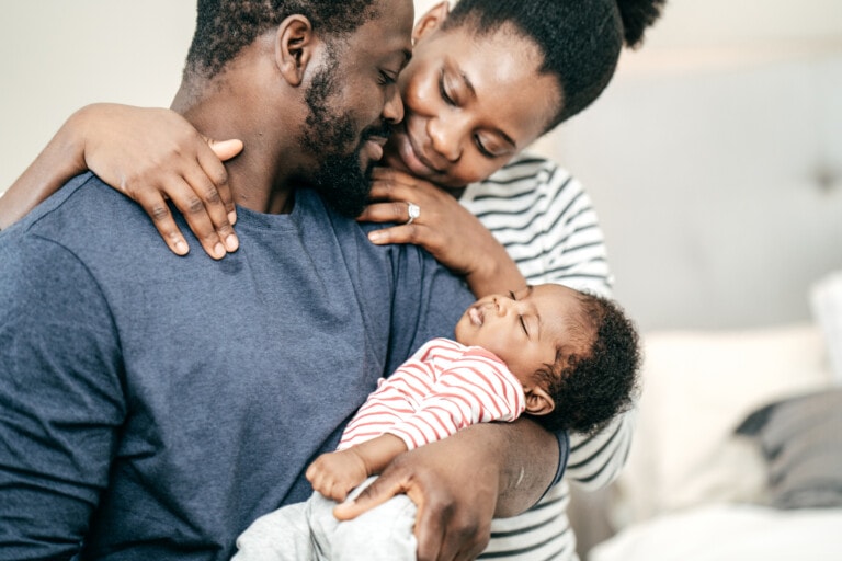A man and woman are sitting close together, embracing while holding a sleeping baby. The man is wearing a blue shirt, and the woman is wearing a striped shirt. The baby in a red and white striped onesie completes this tender scene of little moments captured forever.