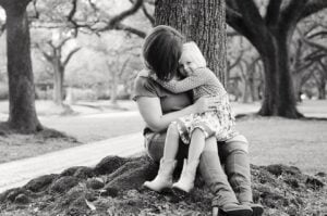 A black-and-white photo of a woman sitting on the ground with her back against a tree, hugging her daughter who is sitting on her lap. The girl has her arms around the woman's neck. Both are in a park with trees in the background, capturing a tender moment that could be from a letter to my daughter.