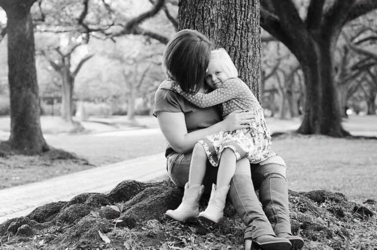 A black-and-white photo of a woman sitting on the ground with her back against a tree, hugging her daughter who is sitting on her lap. The girl has her arms around the woman's neck. Both are in a park with trees in the background, capturing a tender moment that could be from a letter to my daughter.